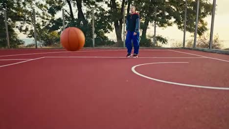 Shooting-close-up-a-blonde-girl-in-a-sports-uniform-picks-up-an-orange-basketball-from-the-red-floor-that-is-bouncing-on-a-street-court-in-the-morning