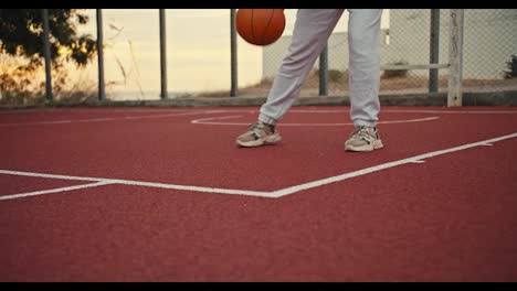 Close-up-shot-of-a-girl-in-white-pants-hitting-an-orange-basketball-on-a-red-basketball-court-early-in-the-morning.-A-girl-in-white-pants-does-exercises-and-practices-handling-an-orange-basketball-in-the-summer