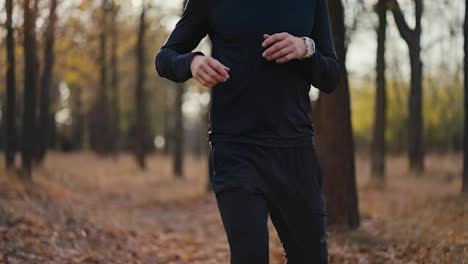 Close-up-footage-of-a-brunette-man-in-a-black-sports-uniform-running-up-to-his-destination-in-the-autumn-forest-and-taking-a-short-break-wiping-away-sweat-during-his-morning-jog-in-a-sunny-autumn-forest