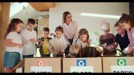 Children,-at-the-command-of-a-teacher,-women-in-a-white-shirt,-put-garbage-into-the-right-boxes-separately,-plastic,-glass-and-paper-in-their-club-for-preparing-children-for-school.-The-woman-teaches-children-how-to-properly-dispose-of-trash