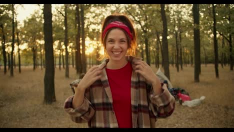 Portrait-of-a-happy-girl-in-a-plaid-shirt-who-straightens-her-hair-and-bandana-and-stands,-smiles-and-rejoices-against-the-background-of-the-rest-of-the-group-of-hikers-and-tents-in-the-green-Sunny-summer-forest