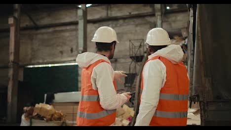 Rear-view-of-a-duo-of-men-in-special-protective-clothing-and-orange-vests-talking-and-discussing-How-to-better-design-plans-at-their-waste-recycling-plant-while-recycling-waste-paper-on-a-conveyor-belt