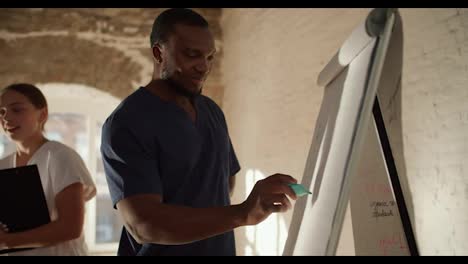 Close-up-shooting:-A-Black-male-doctor-in-a-blue-uniform,-writes-an-explanation-on-a-canvas-with-a-green-felt-tip-pen,-a-nurse-in-a-white-uniform-tells-other-people-information-against-the-background-of-white-brick-walls-in-a-spacious-room