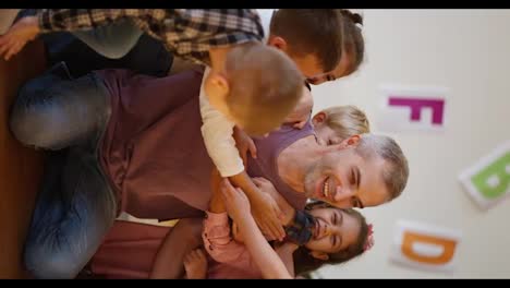 Vertical-video-of-children-running-up-to-their-teacher,-a-man-with-gray-hair-in-a-purple-T-shirt,-and-hugging-him-in-a-club-room-to-prepare-children-for-school