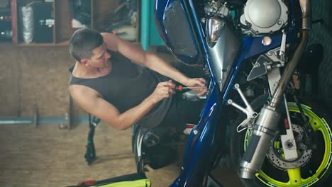 Vertical-video-of-a-male-master-Motor-mechanic-with-a-short-haircut-in-a-gray-T-shirt-repairing-his-blue-and-yellow-bike-and-dismantling-it-into-pieces-in-his-workshop-garage