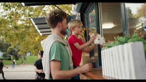 Happy-little-blond-boy-in-a-red-T-shirt-receives-two-hot-dogs-from-the-window-of-a-street-cafe-in-the-park-while-sitting-in-the-arms-of-his-dad-with-curly-hair-in-a-Green-T-shirt.-Father-and-son-having-a-snack-while-walking-in-the-park