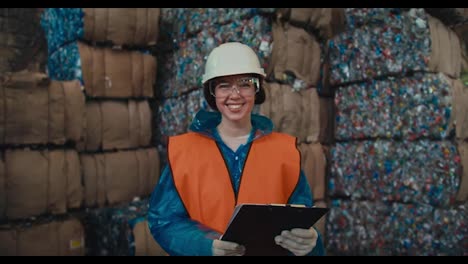 A-brunette-girl-in-a-white-helmet-a-blue-uniform-and-an-orange-protective-vest-holds-a-tablet-in-her-hands-stands-and-looks-at-the-camera-near-a-huge-pile-of-recycled-waste-storage