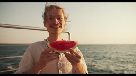 Glücklicher-Blonder-Typ-Mit-Stoppeln-In-Einem-Hellen-Hemd,-Der-Ein-Stück-Wassermelone-In-Seinen-Händen-Hält-Und-Auf-Dem-Pier-In-Der-Nähe-Des-Meeres-Sitzt.-Glücklicher-Typ-Bei-Einem-Picknick,-Der-Ein-Stück-Wassermelone-In-Seinen-Händen-Hält