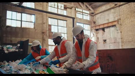 Ein-Trio-Von-Arbeitern-Einer-Abfallrecyclinganlage-In-Speziellen-Schutzuniformen-Und-Weißen-Helmen-Steht-Neben-Dem-Förderband-Und-Legt-Flaschen-Je-Nach-Farbe-Des-Kunststoffs-In-Der-Anlage-Aus.