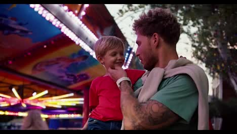 Happy-little-blond-boy-in-a-red-T-shirt-touches-his-dad's-stubble-while-sitting-in-his-arms-in-an-amusement-park.-Dad-and-son-play-while-son-sits-in-dad's-arms-in-amusement-park