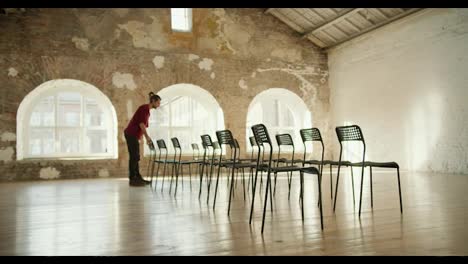 A-brunette-man-with-long-hair-and-a-beard,-wearing-a-red-shirt-and-jeans,-adjusts-black-chairs-in-a-spacious-building-with-white-brick-walls