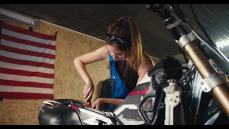 close-up-shot:-a-girl-auto-mechanic-repairs-a-motorcycle-in-her-workshop-with-a-screwdriver-against-the-background-of-the-US-flag-and-tools-in-the-garage