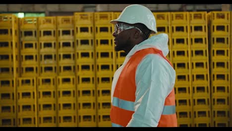 Un-Hombre-De-Piel-Negra-Con-Gafas-De-Seguridad,-Casco-Blanco-Y-Uniforme-Con-Chaleco-Naranja-Camina-Junto-A-Enormes-Estanterías-Con-Cajas-De-Plástico-En-Una-Planta-De-Reciclaje-Y-Clasificación-De-Residuos.