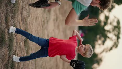 Vertical-video:-A-little-blond-boy-in-a-red-T-shirt-with-a-basketball-glove-throws-a-basketball-into-the-basketball-glove-of-his-dad-in-a-Green-T-shirt-on-the-court-in-the-park