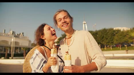 Portrait:-Happy-couple-on-a-date-on-a-modern-beach,-a-blond-guy-with-stubble-in-a-light-shirt-holds-a-hot-dog-along-with-his-girlfriend-who-also-holds-a-hot-dog-and-poses-and-looks-at-the-camera-on-a-date-near-the-sea