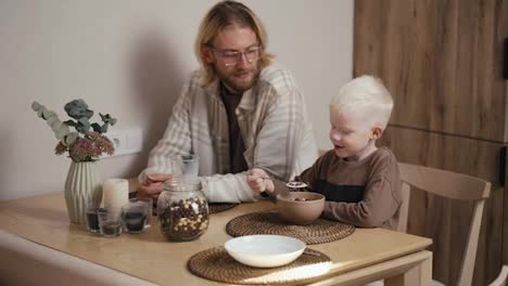 Glücklicher-Kleiner-Albino-Junge-Mit-Weißer-Haarfarbe-In-Einer-Braunen-Jacke-Isst-Müsli-Mit-Milch-Und-Frühstückt-Morgens-Mit-Seinem-Blonden-Vater-Mit-Bart-Und-Brille-In-Einer-Modernen-Küche