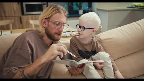 Happy-father,-a-blond-man-in-glasses-with-a-beard-explains-to-his-little-son,-an-albino-boy-with-white-hair-color-in-blue-glasses,-how-to-read-and-complete-an-assignment-in-elementary-school-during-homework-at-home-in-the-kitchen