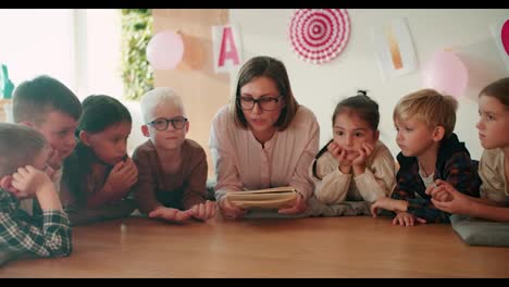 Una-Niña-Rubia-Con-Un-Peinado-Bob-Con-Gafas-Y-Una-Camisa-Blanca-Lee-Un-Libro-A-Niños-En-Edad-Preescolar-Mientras-Decide-Sentarse-En-El-Suelo-Sobre-Almohadas-Especiales-Con-Los-Niños-Que-La-Escuchan.