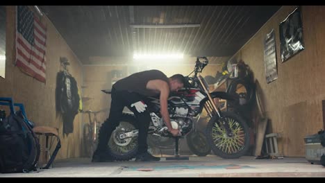 A-male-mechanic-with-a-short-haircut-in-a-gray-T-shirt-removes-his-bike-from-the-jack-and-takes-it-out-of-his-garage-workshop.-Male-mechanic-completing-motorcycle-repair