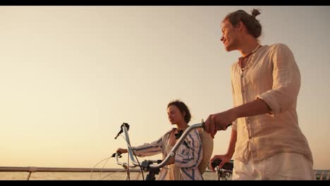 Happy-couple-guy-and-girl-walking-together-with-their-bicycles-along-the-beach-near-the-sea-at-sunrise.-A-guy-in-light-beachwear-and-a-girl-in-a-blue-white-shirt-walk-with-their-bicycles-along-the-beach-against-the-sky-at-Sunrise