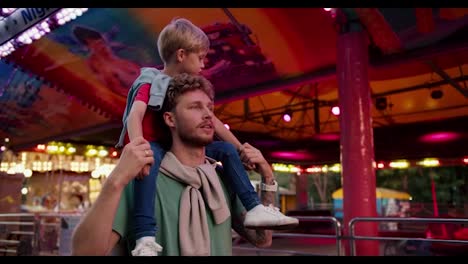 A-happy-dad-with-curly-stubble-hair-in-a-Green-T-shirt-carries-his-little-blond-son-in-a-red-T-shirt-on-his-shoulders-and-they-look-around-in-the-amusement-park.-Bright-lights-and-interesting-images-capture-the-eye-and-interest-the-little-blond-boy-who-sits-on-his-dad's-shoulders