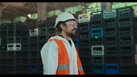 A-brunette-man-with-a-beard-in-a-protective-suit-goggles-and-a-helmet-in-an-orange-vest-walks-along-black-and-yellow-boxes-for-sorting-and-recycling-waste-at-a-large-factory