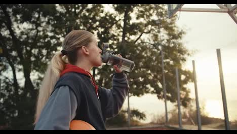 Nahaufnahme-Der-Rückansicht-Eines-Blonden-Mädchens-Mit-Pferdeschwanzfrisur,-Das-Während-Ihres-Morgendlichen-Basketballtrainings-Wasser-Aus-Einer-Sportflasche-Trinkt-Und-Den-Sonnenaufgang-Im-Sommer-Betrachtet