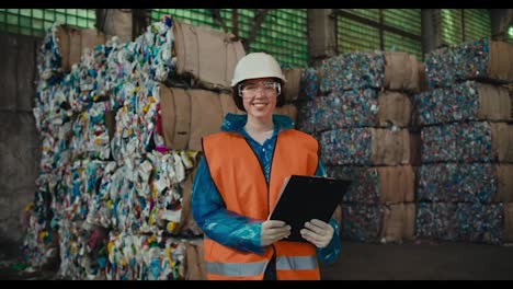 Portrait-of-a-happy-brunette-girl-in-a-blue-protective-uniform-and-an-orange-vest-who-is-holding-a-tablet-in-her-hands-smiling-and-looking-at-the-camera-near-a-large-pile-of-recycled-plastic-at-a-waste-recycling-plant