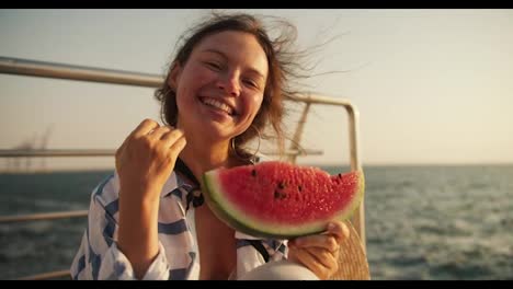 Portrait-of-a-happy-girl-who-smiles-and-looks-at-the-camera-and-holds-a-slice-of-watermelon-in-her-hands-on-a-pier-near-the-sea.-A-brown-haired-girl-smiles-and-laughs-cheerfully-and-holds-a-slice-of-watermelon-in-her-hands-on-a-pier-near-the-sea