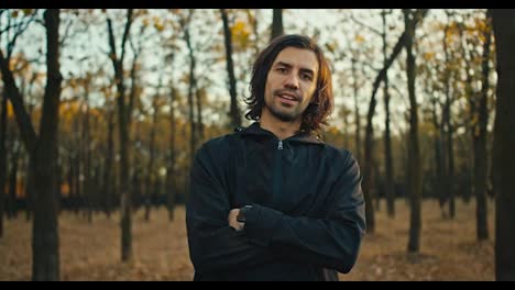 Confident-serious-man-with-medium-length-hair-and-a-brunette-beard-holds-his-arms-crossed-on-his-chest-in-a-black-sports-uniform-stands-in-the-autumn-forest