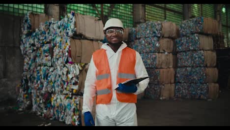 Portrait-of-a-happy-man-with-Black-skin-in-a-white-uniform-and-an-orange-vest-who-stands-near-a-huge-pile-of-recycled-plastic-at-a-waste-recycling-plant-and-holds-a-tablet-in-his-hands