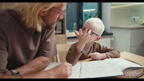 Un-Niño-Albino-Interesado-Con-Gafas-Azules-Y-Cabello-Blanco-Se-Comunica-Con-Su-Padre,-Un-Hombre-Rubio-Con-Barba-Y-Gafas,-Sobre-Un-Tema-Determinado-En-La-Escuela-Y-El-Niño-Quiere-Aprender-Cosas-Nuevas-En-La-Cocina-En-Un-Apartamento-Moderno-Con-Su-Padre.