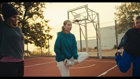 Un-Trío-De-Chicas-En-Ropa-Deportiva-Calentándose-Antes-De-Jugar-Al-Baloncesto-En-El-Patio-De-Recreo-Rojo-Cercado-Por-Una-Valla-Cerca-De-Una-Pelota-De-Baloncesto-Naranja-En-Una-Mañana-De-Verano.