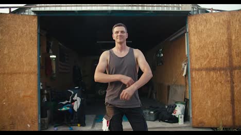 A-happy-male-mechanic-in-a-gray-T-shirt-has-his-hands-folded-on-his-chest,-standing-against-the-backdrop-of-an-open-garage-workshop.-Portrait-of-a-mechanic-near-his-workshop