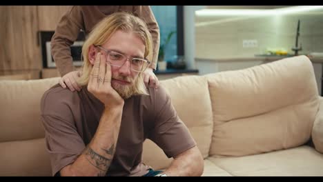 A-tortured-blond-man-with-a-beard-and-glasses-sits-on-the-sofa-while-his-little-albino-son-pesters-him-and-wants-to-play-with-the-man-at-home-in-a-modern-apartment.-Tired-man-with-glasses-cannot-pay-enough-attention-to-his-little-albino-son-in-a-modern-apartment