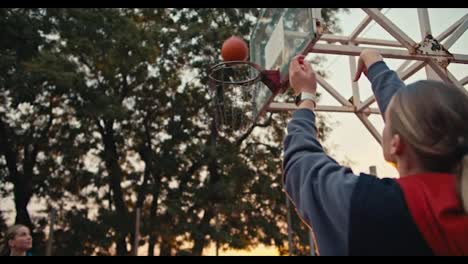 Close-up-shot-of-a-blonde-girl-in-a-hoodie-throwing-a-basketball-into-the-basket-while-playing-with-her-friend-on-a-fenced-in-basketball-court-at-Sunrise-in-the-morning-in-summer