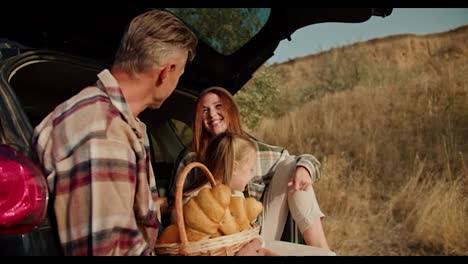 A-man-with-gray-brunette-hair-communicates-with-his-happy-wife-in-a-green-checkered-shirt-and-their-little-daughter-a-blonde-girl-while-they-sit-in-an-open-trunk-during-their-picnic-outside-the-city-in-the-summer