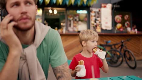 A-little-blond-boy-with-blue-hair-in-a-red-T-shirt-eats-hot-dogs-at-a-street-table-in-a-street-cafe-while-his-business-dad-in-a-Green-T-shirt-with-stubble-talks-on-the-phone.-The-problem-of-lack-of-attention-for-young-children-from-their-parents