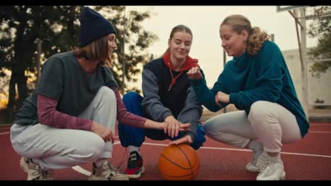 A-trio-of-happy-blonde-girls-on-the-same-basketball-team-put-their-hands-together-and-raise-them-up-in-a-sign-of-unity-near-the-basketball-ball-on-the-red-basketball-court-early-in-the-morning