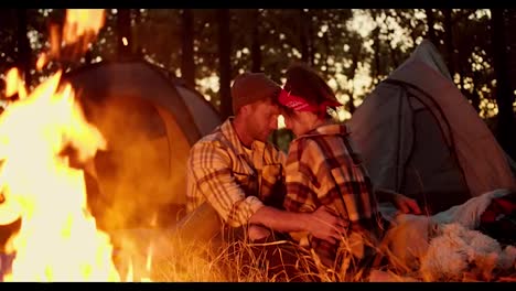 Romantic-evening,-a-man-in-a-checkered-shirt-and-a-hat-sits-near-a-girl-with-a-red-bandana-and-in-a-checkered-shirt-near-a-fire-against-the-backdrop-of-tents-and-they-closely-communicate-with-each-other-about-something-against-of-a-forest-in-the-fall