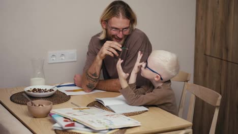 Happy-blond-man-with-a-beard-and-glasses-helps-his-little-albino-son-with-white-blond-hair-in-blue-glasses-to-count-a-math-assignment-using-his-fingers-on-his-hand-and-do-his-homework-while-preparing-for-the-next-day-at-school-in-a-modern-kitchen-at-home