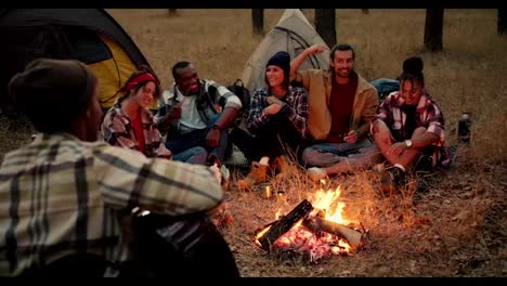 Un-Hombre-Con-Sombrero-Toca-La-Guitarra-Y-El-Resto-Del-Grupo-De-Excursionistas-Toca-Con-él-Y-Se-Divierte-Y-Canta-Mientras-Está-Sentado-Cerca-De-Un-Pequeño-Fuego-En-El-Bosque.-Un-Grupo-Alegre-Y-Feliz-De-Participantes-De-Una-Caminata-Se-Sienta-Cerca-Del-Fuego-Y-Se-Divierte-Con-Canciones-Y-Guitarra.