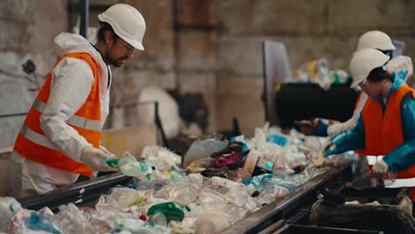 A-man-in-a-white-uniform-an-orange-vest-and-a-white-helmet-together-with-two-of-his-colleagues-sorts-plastic-bottles-near-a-conveyor-belt-at-a-large-waste-recycling-and-sorting-plant