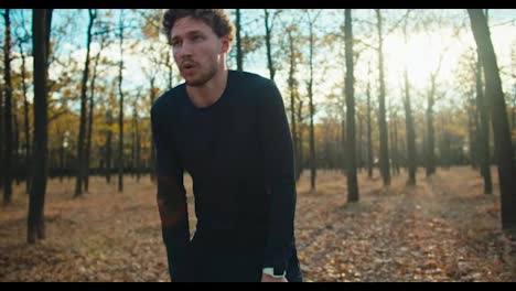 A-tired-man-with-curly-hair-in-a-black-sports-uniform-runs-to-his-destination-wipes-sweat-from-his-forehead-and-looks-at-the-time-of-his-run-in-the-morning-in-the-autumn-forest-on-a-sunny-morning