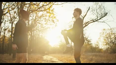 A-man-with-curly-hair-in-a-black-sports-uniform-together-with-his-friend-in-a-red-cap-and-black-shorts-are-warming-up-before-their-jogging-in-the-autumn-park-at-Sunrise-on-a-sunny-morning-in-autumn