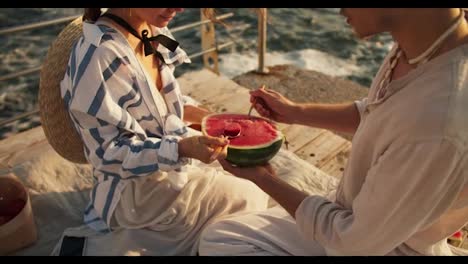 A-guy-and-a-girl-on-a-picnic-on-a-pier-near-the-sea-eat-watermelon-with-spoons.-Happy-couple,-a-blond-guy-with-tied-hair-or-a-brown-haired-girl-in-a-white-blue-shirt-are-sitting-on-a-pier-near-the-sea-and-eating-watermelon