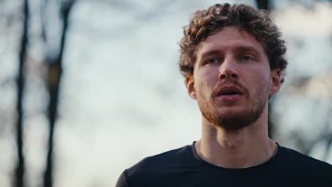 Porträt-Eines-Mannes-Mit-Lockigem-Haar-Und-Bart-In-Schwarzer-Sportuniform,-Der-Sich-Nach-Einem-Morgendlichen-Lauf-Im-Wald-Den-Schweiß-Aus-Dem-Gesicht-Wischt