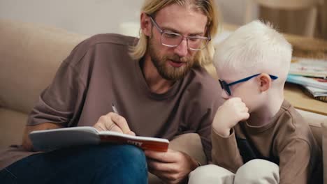 A-blond-man-in-glasses-with-a-beard-helps-his-little-son,-an-albino-boy-in-glasses-with-white-hair,-to-sort-out-his-homework-assignment-and-read-the-sentence-correctly.-Blond-man-doing-homework-with-his-little-albino-boy