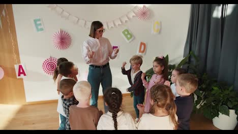 Happy-preschool-children-in-a-group-doing-a-wrist-warm-up-before-writing-with-their-teacher,-a-girl-with-a-bob-hairstyle-and-glasses-in-a-white-shirt-in-a-special-club-for-preparing-children-for-school