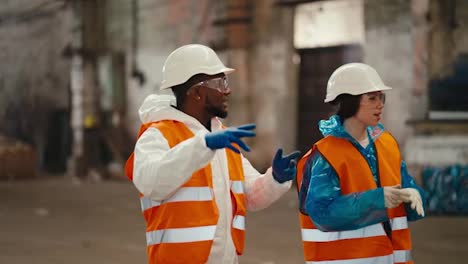 A-man-with-Black-skin-in-a-white-uniform-together-with-his-colleague-a-girl-in-an-orange-vest-walk-along-the-large-hall-of-a-waste-processing-plant-and-communicate-with-each-other